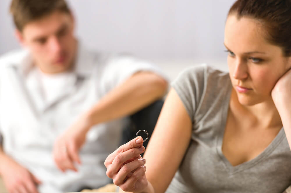 A disagreeing couple sit on a couch and one holds up their wedding ring